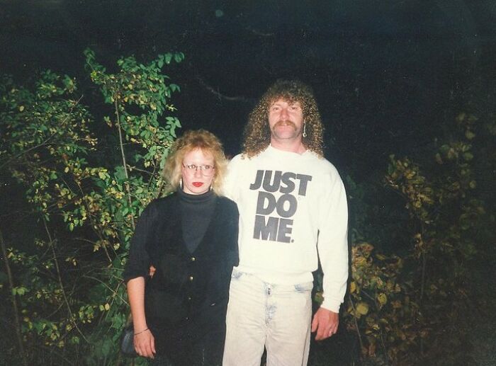 Funny couple posing outdoors, woman in black and man with curly hair in "Just Do Me" shirt, night background.