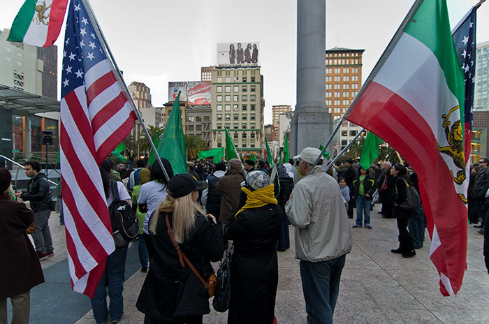 Crowd gathered at a public square holding American and historical Iranian flags, illustrating real proven conspiracies.