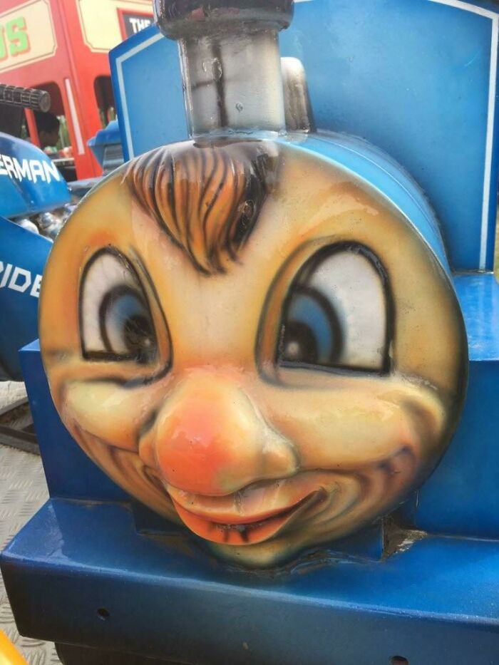 Face of a hilariously creepy toy train with exaggerated features and a sinister smile on a blue ride vehicle.