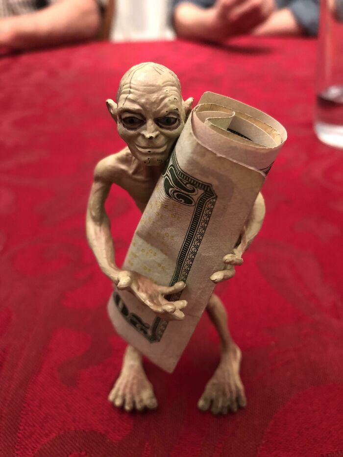 Creepy toy figurine resembling Gollum holding a rolled-up dollar bill on a red patterned surface.