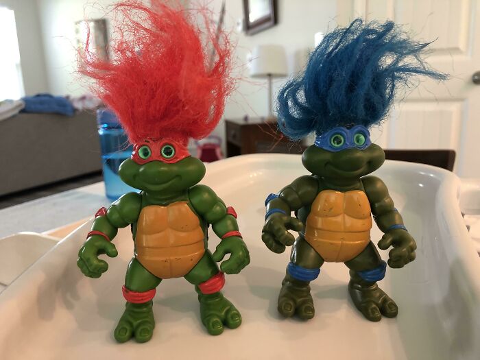 Two creepy toys resembling turtles with bright red and blue wild hair standing on a white surface indoors.