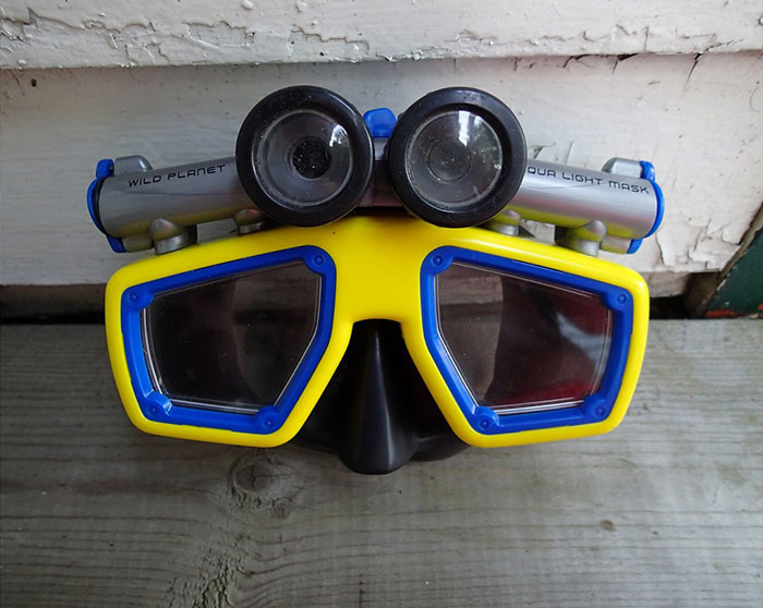 Yellow and blue deep sea diving mask with attached underwater lights resting on wooden surface.