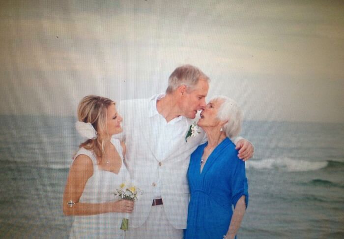 Couple sharing a humorous moment by the beach with an elderly woman, dressed in wedding attire, evoking laughter.