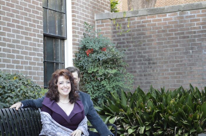 Smiling couple posing on a bench in a garden setting, with the woman in a purple dress, capturing a funny moment.