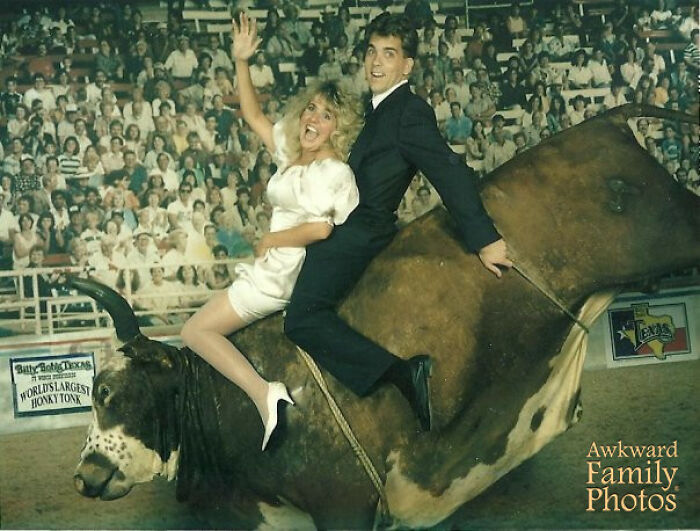 Funny couple riding a fake bull in front of a large crowd, with the woman waving and laughing.