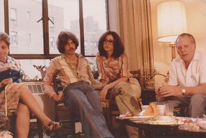 Couple with 70s fashion vibe, sitting casually in a living room, surrounded by family, creating a humorous and nostalgic scene.