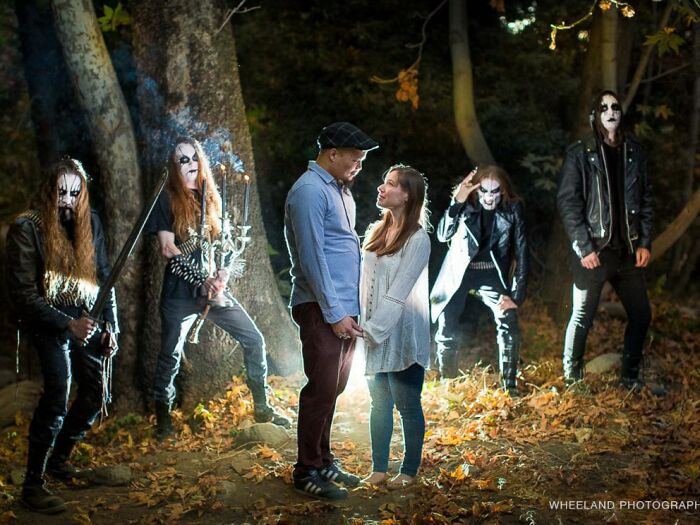 Couple posing romantically in a forest, surrounded by costumed metal band members, creating a funny contrast.