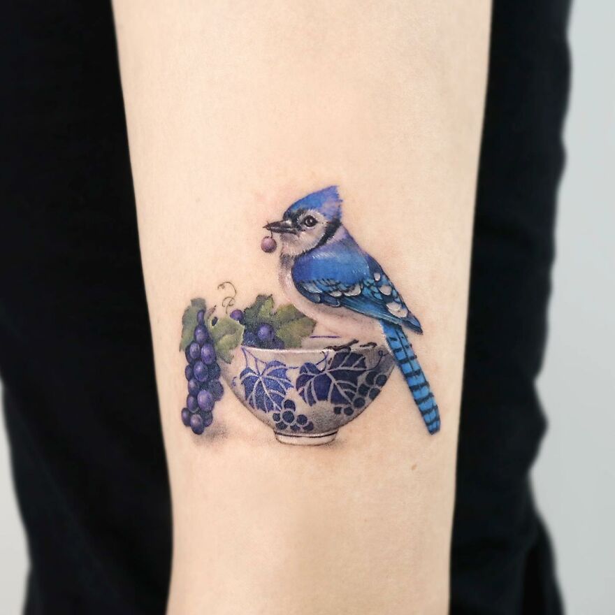 Painting-like tattoo of a blue jay perched on a floral bowl with grapes on an arm.