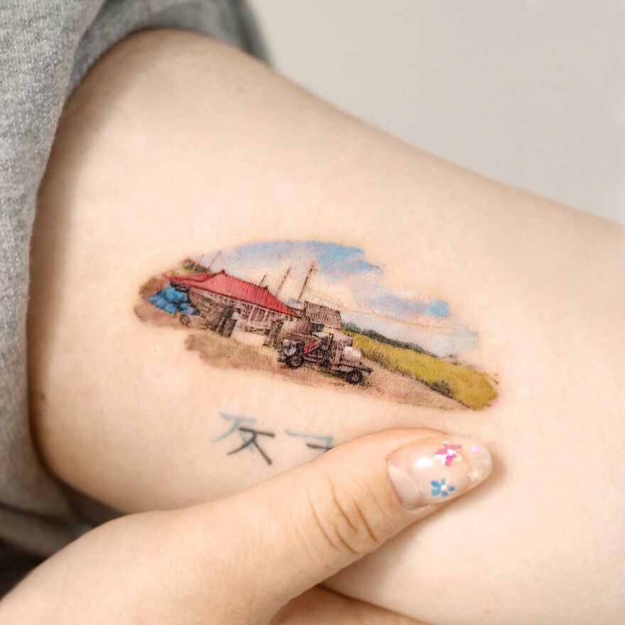 Painting-like tattoo of a rural scene with buildings on arm, vivid colors and detailed artistry.