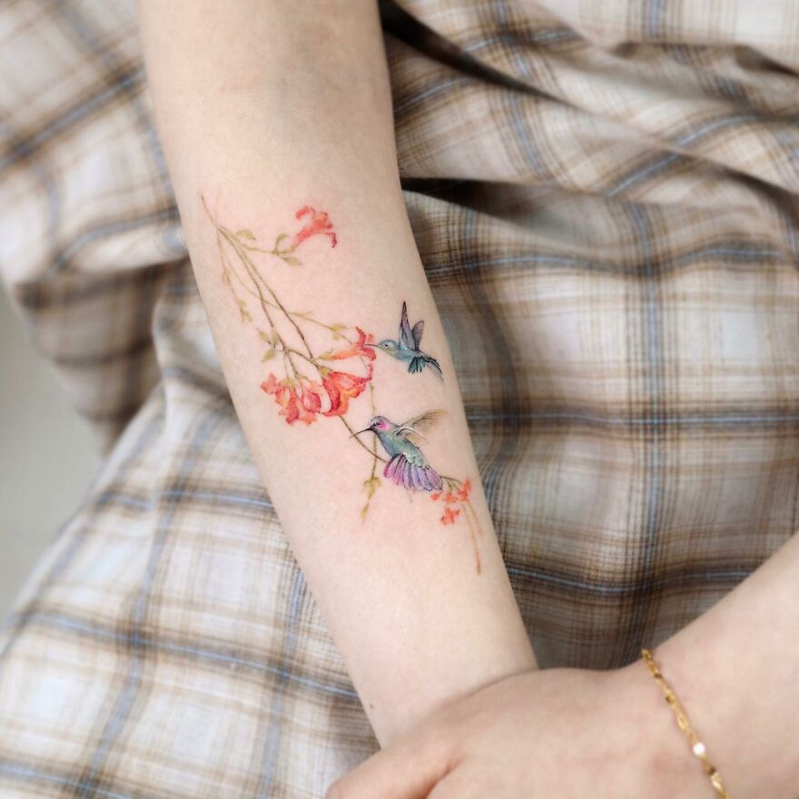 Painting-like tattoo of colorful hummingbirds and flowers on a person's forearm.
