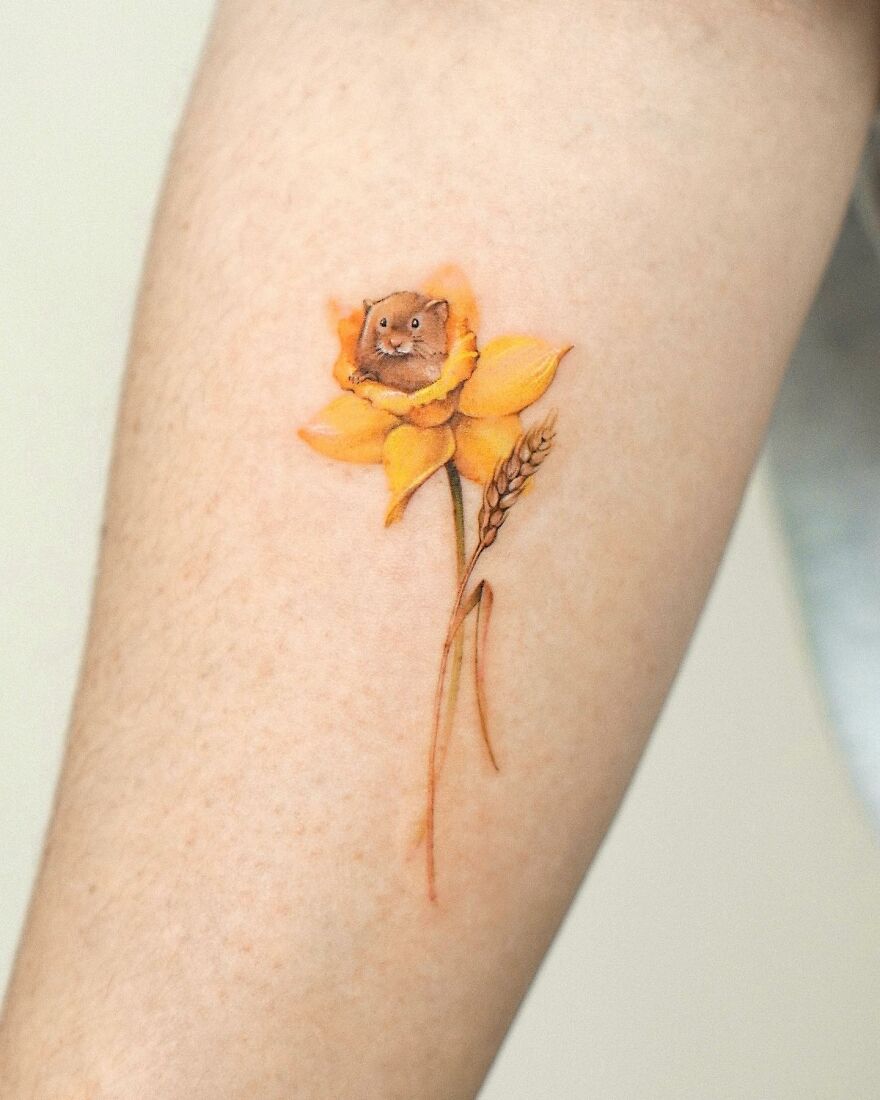 Tattoo of a mouse inside a yellow flower on skin, showcasing painting-like detail by the artist.
