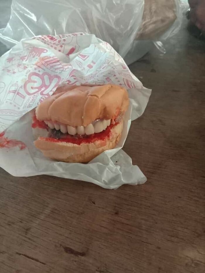 Cursed food burger with fake teeth as the filling, wrapped in branded paper on a wooden table.