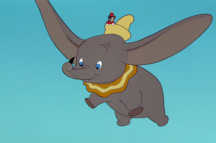 Cute animated elephant with large ears flying, wearing a yellow hat, followed by a tiny mouse, in a clear blue sky.