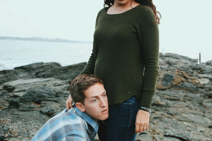 A couple posing humorously on rocky shore, man resting his head on woman's waist, creating a funny moment.