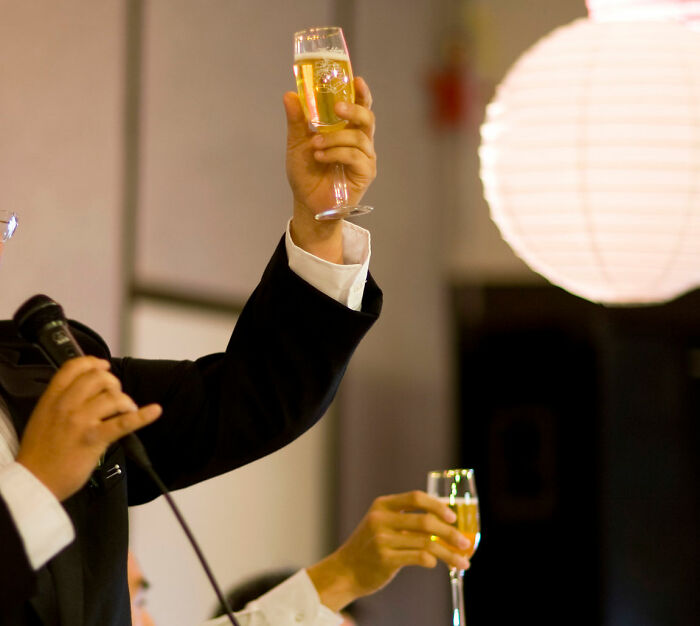Man in a suit holding a microphone and raising a glass during a wedding speech, capturing a moment of a horrible speech.