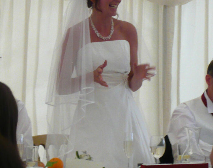 Bride in a wedding dress looking uneasy while giving a speech during a wedding reception with guests around the table.