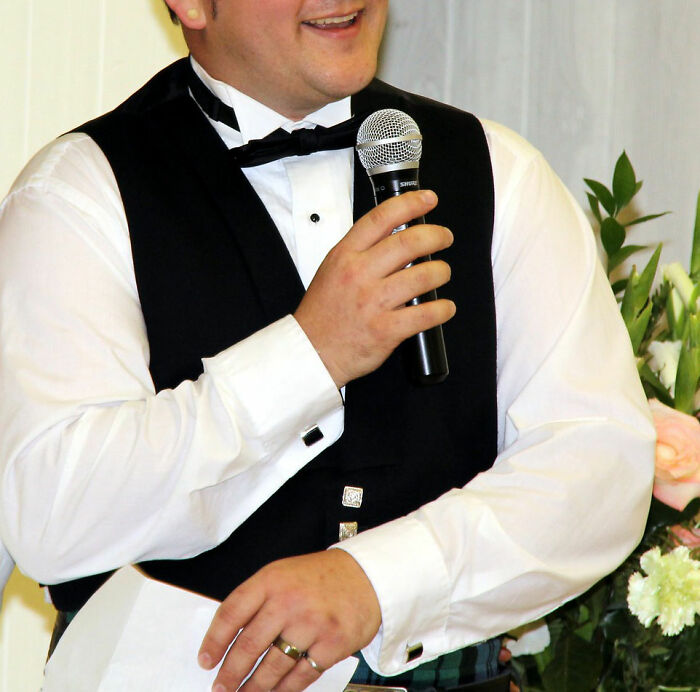 Man in formalwear holding a microphone and paper, giving a wedding speech that causes discomfort among guests.