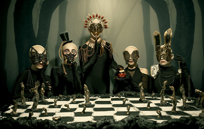 Dark Alice theme with four masked figures in elaborate costumes gathered around a chessboard.