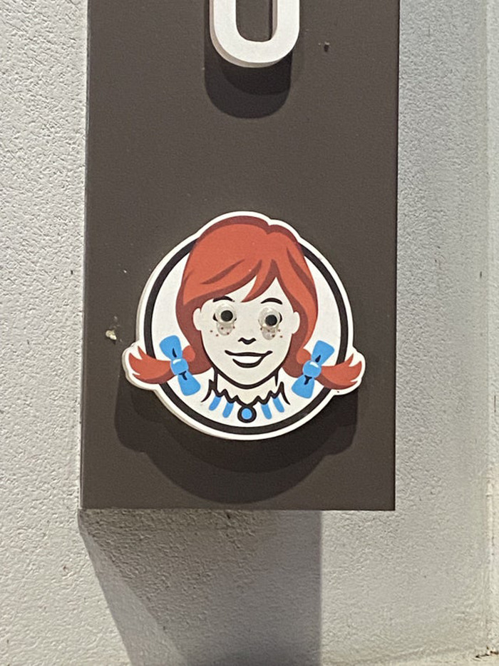 Sticker with wiggle eyes added to a character's face on a wall, enhancing the image with eyebombing creativity.