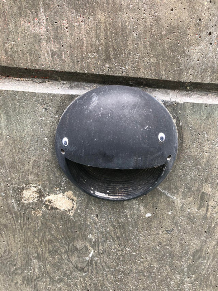Black round vent on concrete wall with wiggle eyes creating a smiling face, showcasing eyebombing making objects better.