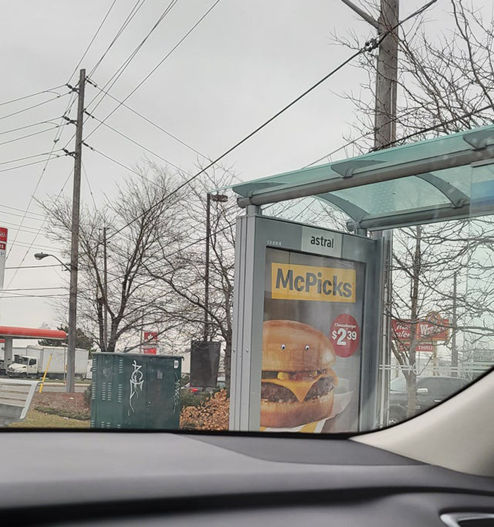 Bus stop ad for a cheeseburger with wiggle eyes added, creating a humorous eyebombing effect on the random object.