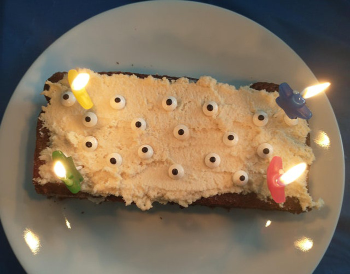 A rectangular cake with cream frosting decorated with multiple wiggle eyes and four lit candles on a white plate.