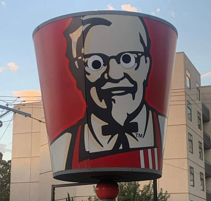 Large KFC bucket sign with wiggle eyes added to Colonel Sanders' face, enhancing the object with eyebombing creativity outdoors.