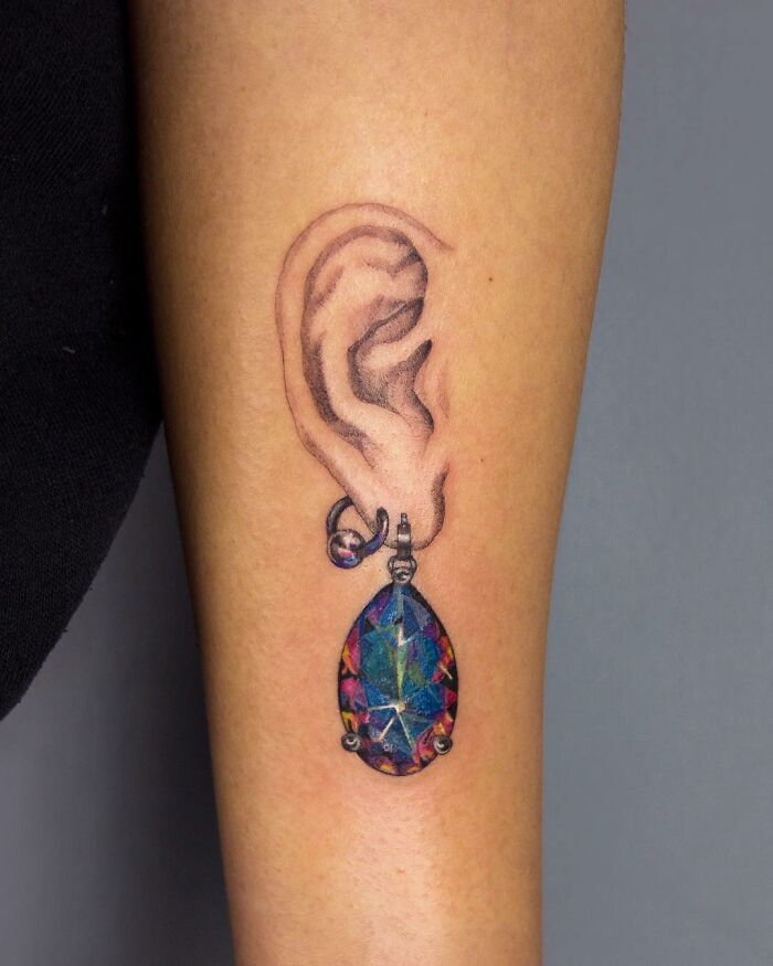 Tattoo of an ear with colorful gemstone earring, showcasing permanent jewelry art.