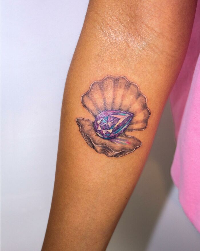 Tattoo of a vibrant gemstone in an open clam shell on a person's arm, showcasing permanent jewelry art.