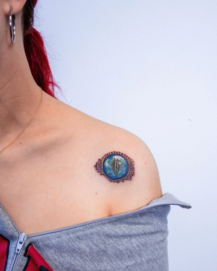 Close-up of a person's shoulder displaying a "permanent jewelry" tattoo, resembling an intricate, colorful brooch.