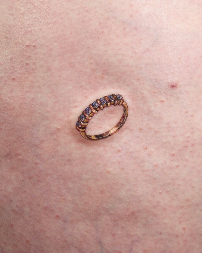 Tattoo of a detailed permanent jewelry ring on skin.