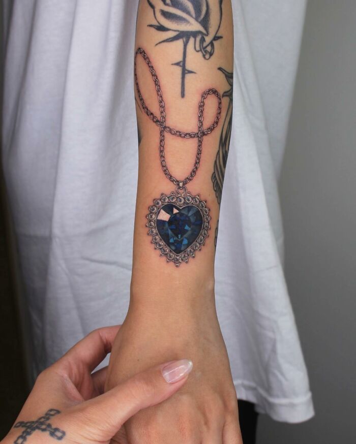 Tattoo of a heart-shaped pendant with chain on an arm, depicting a "permanent jewelry" style.