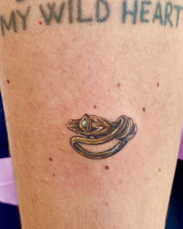 Detailed jewelry tattoo resembling a gold ring on skin with text above.