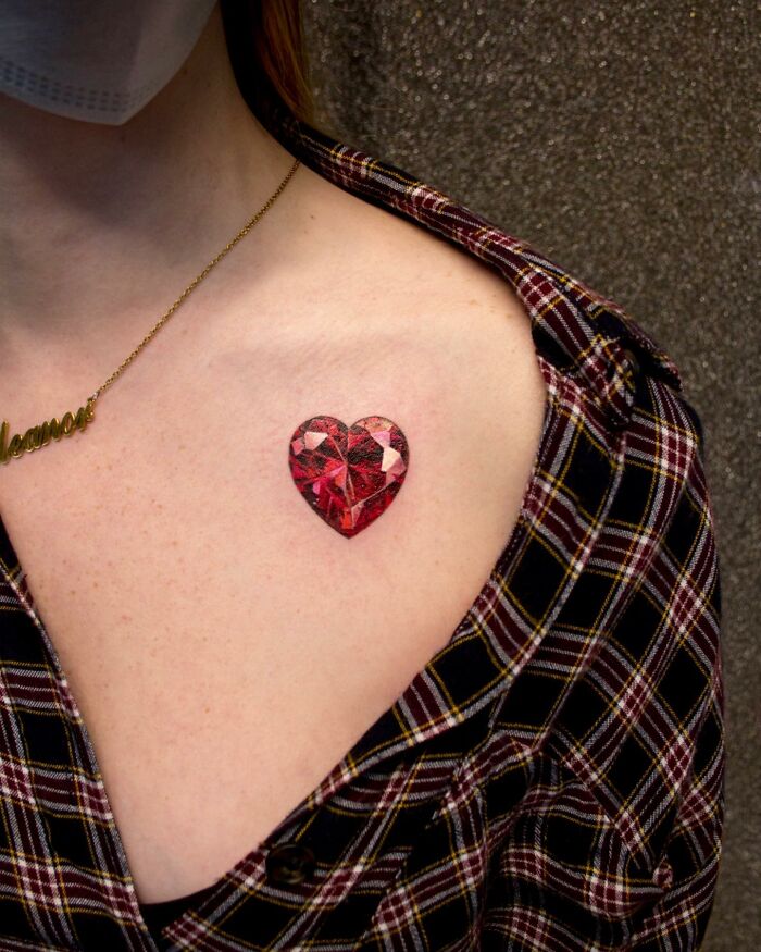Close-up of a "permanent jewelry" tattoo featuring a heart-shaped gem on a person's shoulder.