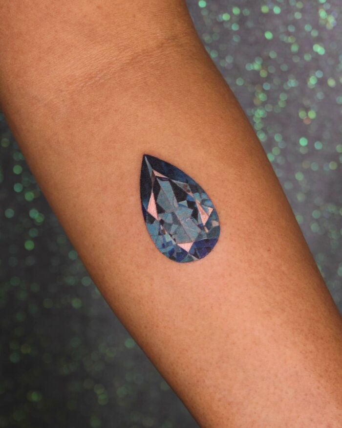 A colorful teardrop gem tattoo on forearm representing permanent jewelry tattoos, with a glittery background.