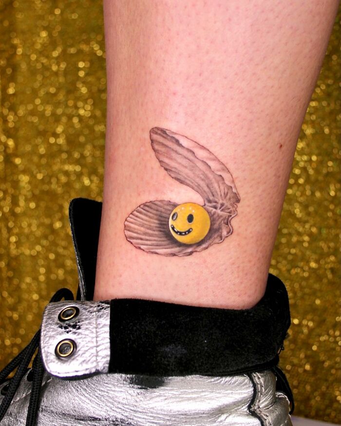 Tattoo of a smiling pearl in a clam shell, symbolizing permanent jewelry art on skin, against a gold background.