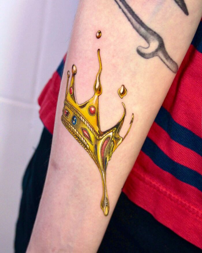 Tattoo of a melting gold crown, symbolizing permanent jewelry, on a forearm wearing a striped shirt.