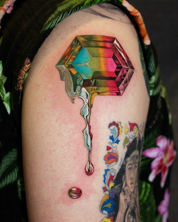 Colorful gem tattoo resembling permanent jewelry on upper arm.