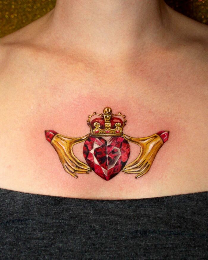 Detailed permanent jewelry tattoo of a crowned heart with golden hands on someone's chest.