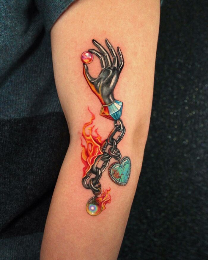 Arm with colorful permanent jewelry tattoo of a chain, flames, and a hand holding a gem.