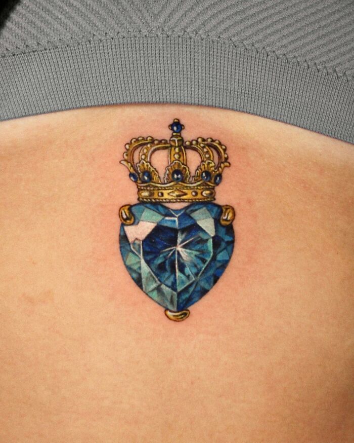 Close-up of a permanent jewelry tattoo featuring a crowned blue gemstone on skin.