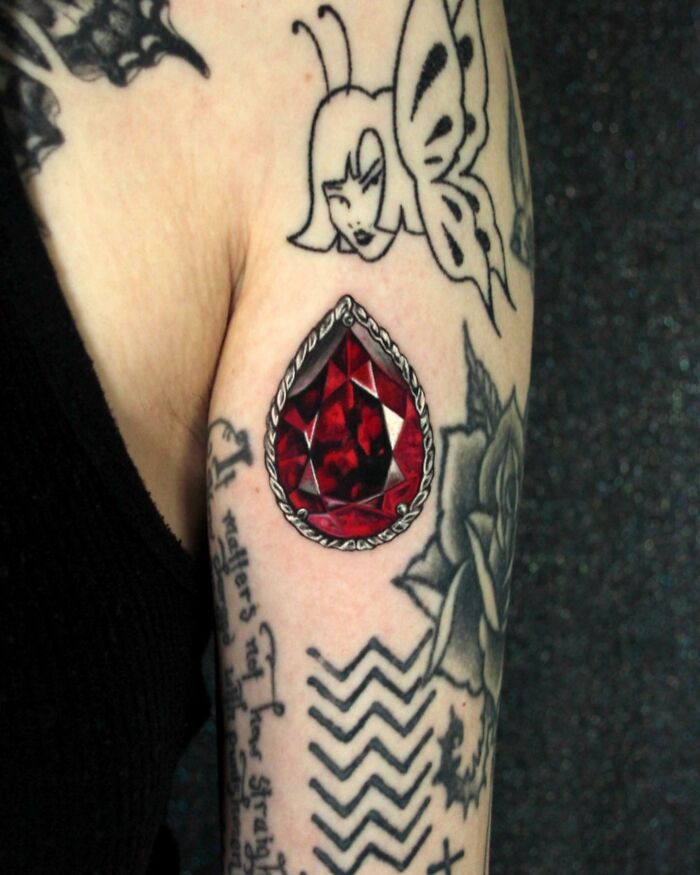 Arm with detailed permanent jewelry tattoos, featuring a red gem and other intricate designs.