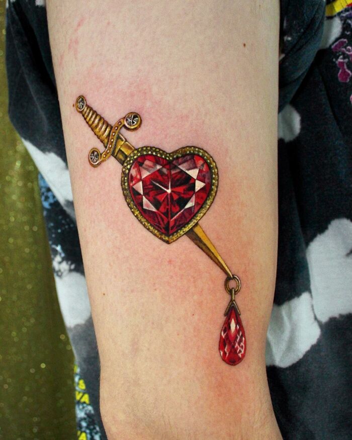 A realistic "permanent jewelry" tattoo of a jeweled heart pierced by a sword with a teardrop gem accent, on a person's arm.