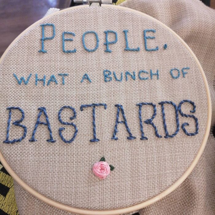 Embroidery hoop with dark humor text: “People, what a bunch of bastards,” featuring a small pink rose.