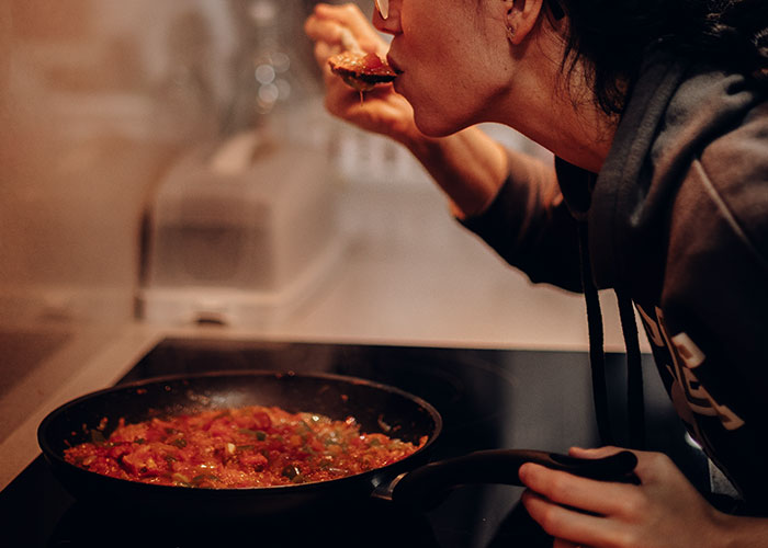 Person secretly tasting food from a pan on the stove while cooking in their kitchen.