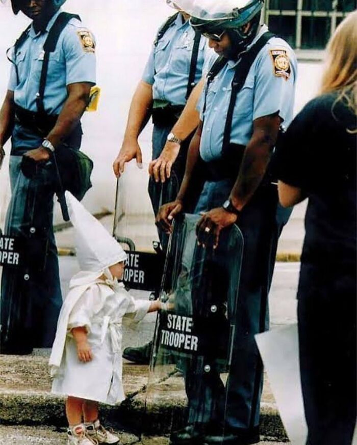 ‘No One Is Born Racist’ Photograph By Todd Robertson. A Boy Dressed In A Kkk Robe And Hood, Curiously Touches The Shield Of A State Trooper During A Kkk Rally | Gainesville, Georgia, 1992