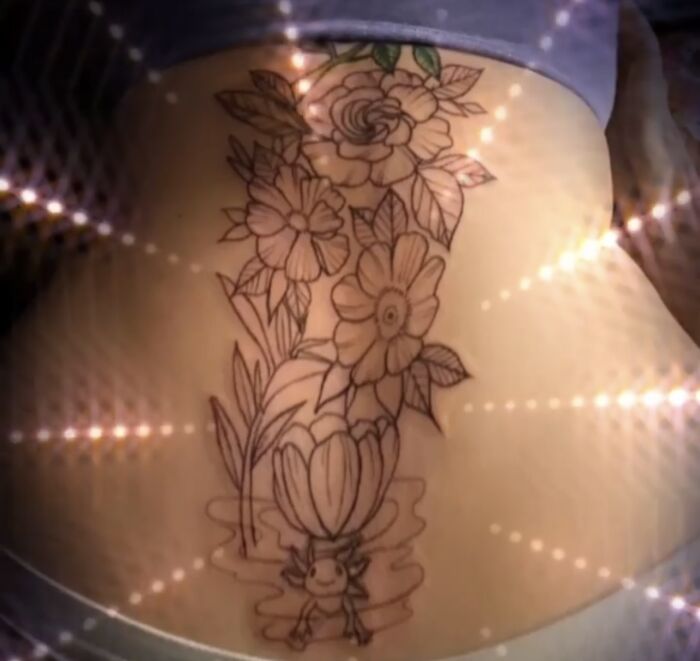 Flower tattoo design on lower back, featuring various blooms and a small character at the base.