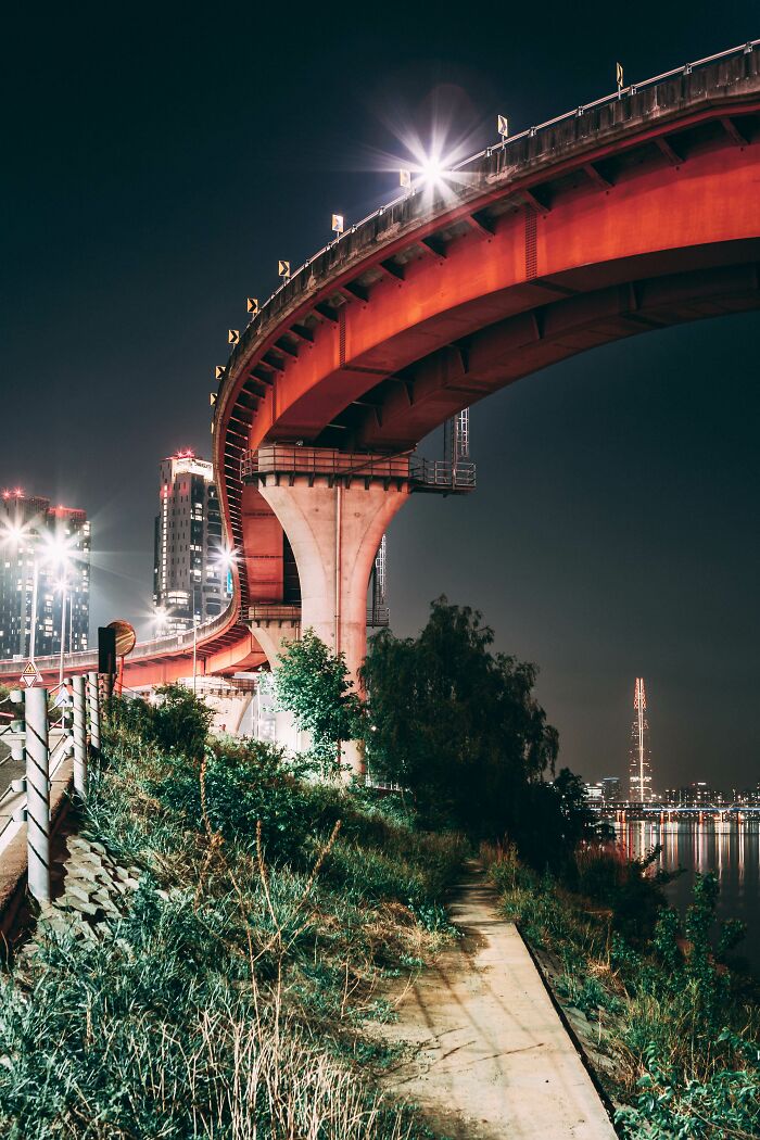 Curved elevated highway lit at night over grass path with Seoul cityscape in the background, showcasing Seoul's beauty.