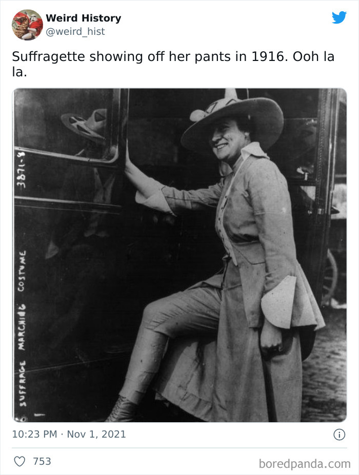 Black and white photo of a smiling suffragette in 1916 wearing pants and a wide-brimmed hat beside a vehicle.