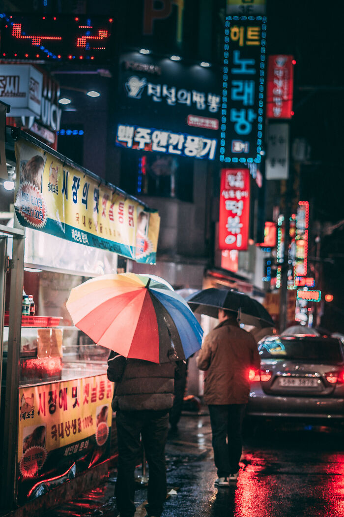 Night street scene in Seoul with colorful umbrellas and bright neon signs reflecting on wet pavement.